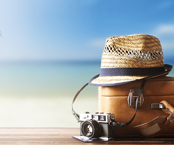 Foto de uma câmera fotográfica, uma mala de viagens e um chapéu com a praia ao fundo.
